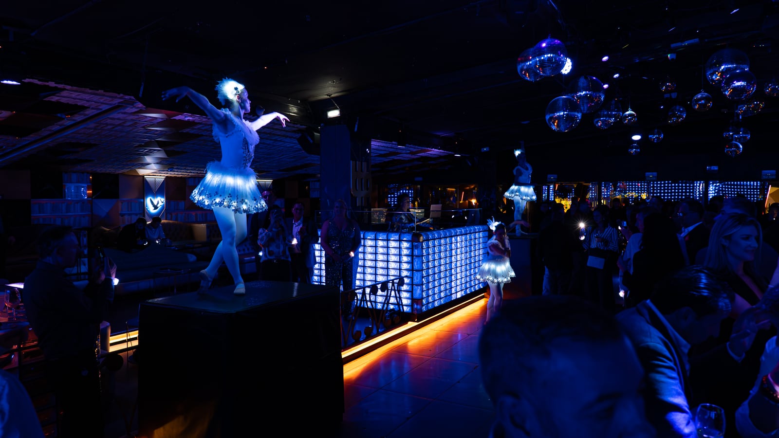 An aerial ballet performer in a light-up costume above a blue-lit bar.