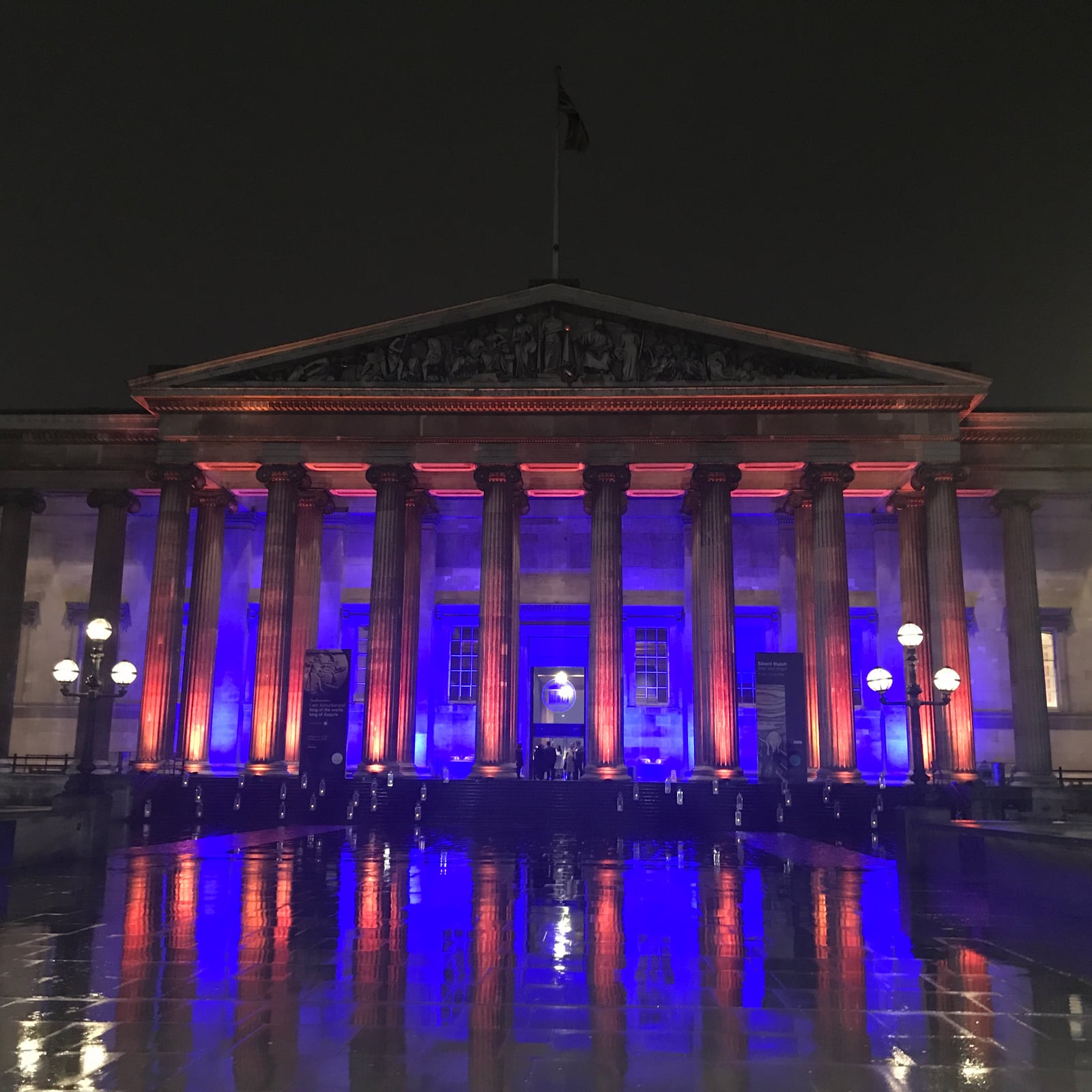 The British Museum's portico lit in purple and blue on a wet evening, reflected in the rain.