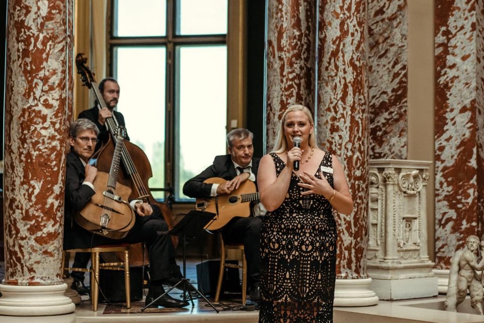 Jennifer with a microphone alongside a guitar and double-bass trio in a marble gallery at Chantilly.