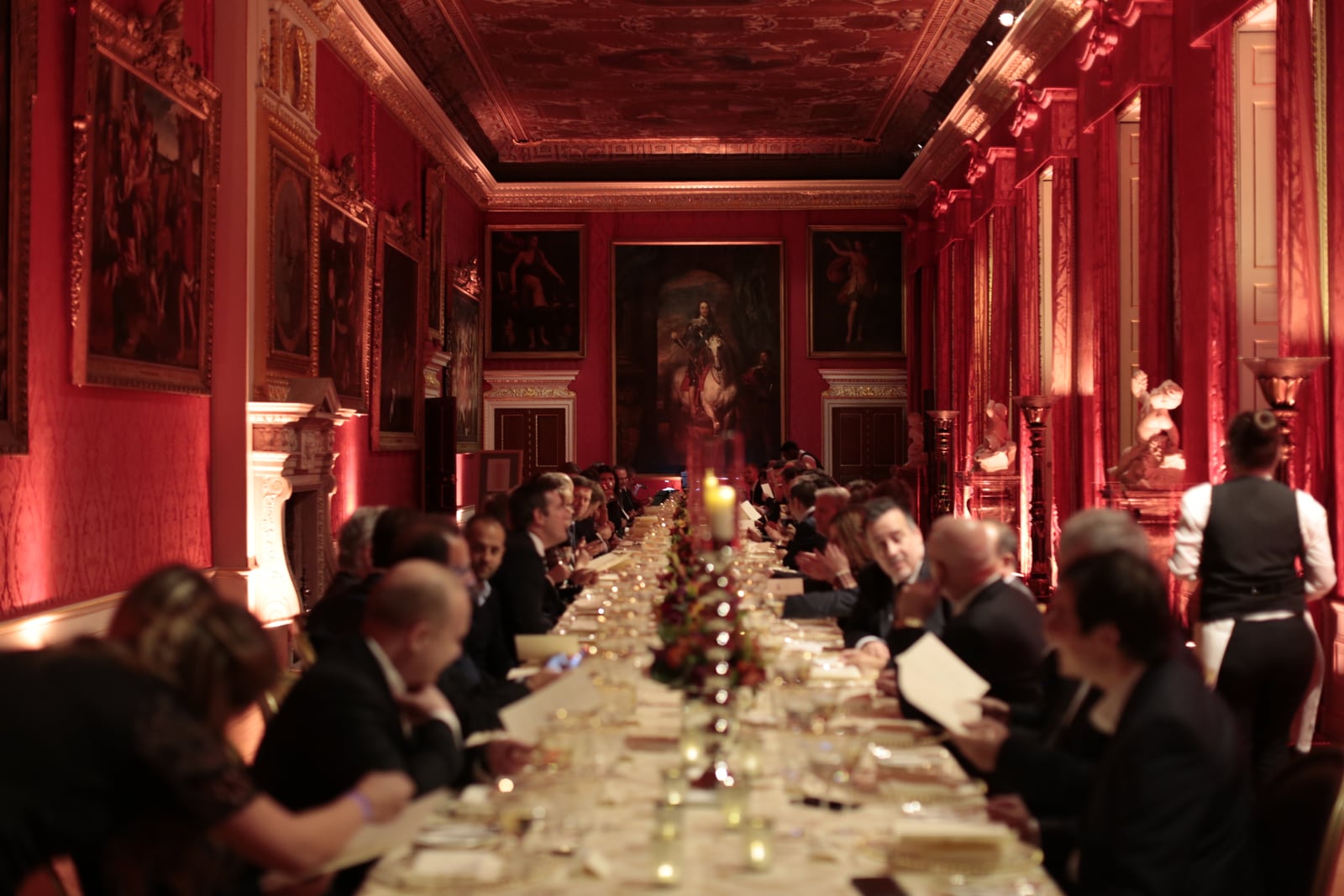 A candlelit long table laid for dinner in the red-walled King's Gallery at Kensington Palace.