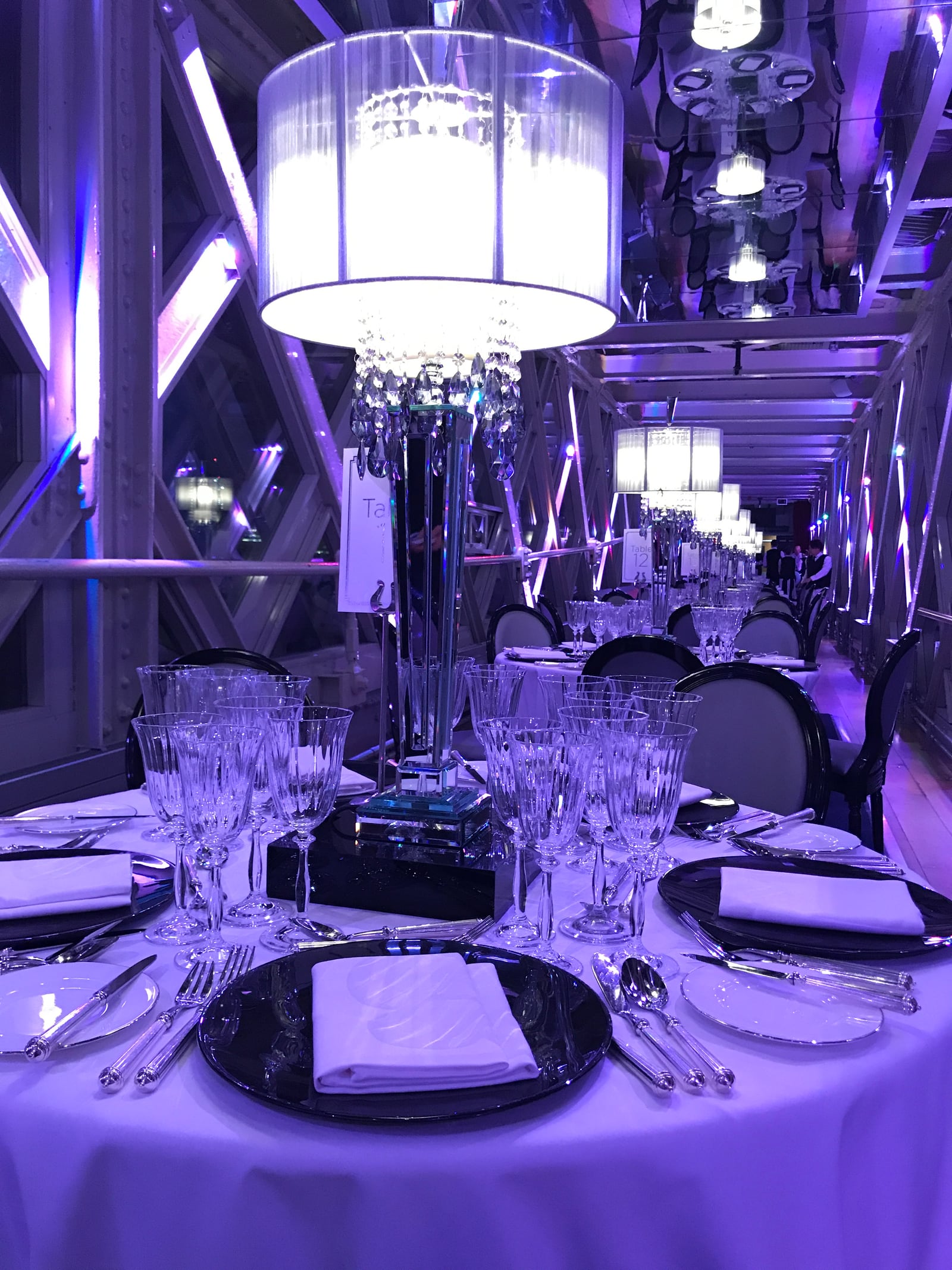 A dinner table with crystal glassware and a table-top chandelier inside the purple-lit upper walkway of Tower Bridge.
